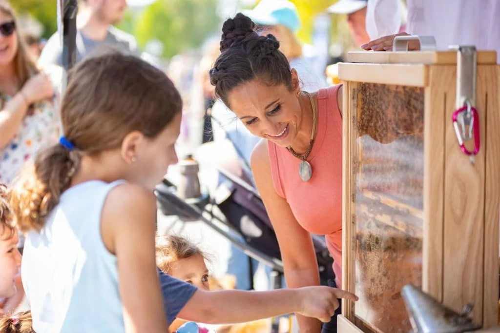 Woman and child interacting with a wooden observation hive at an outdoor event.